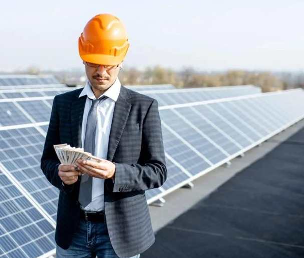 In Jaipur, Rajasthan, a man in a suit and hardhat presents money in front of solar panels, representing solar energy investments